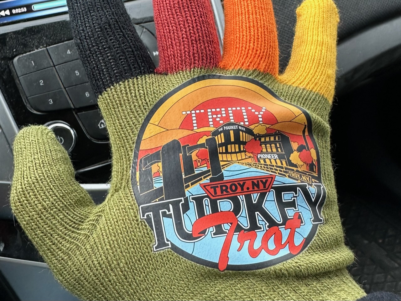 The back of a hand in a multicolored knit glove. Most of the glove is an olive green. The index finger is black, middle finger is red, ring finger is orange, and the pinkie is yellow. On the back of the glove is a graphic which reads "Troy, NY Turkey Trot".