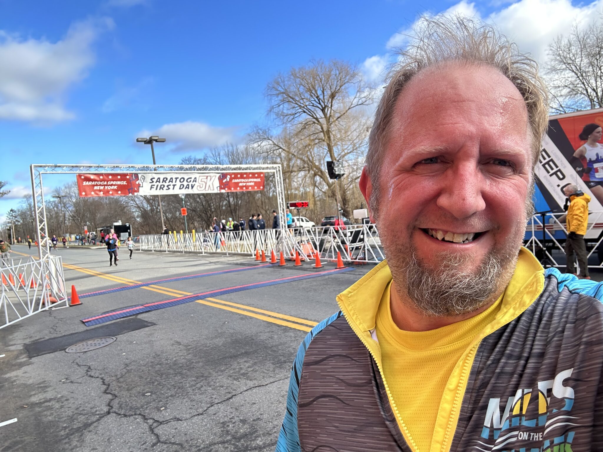 Smiling man with red face and a goatee wearing athletic clothing. In the background is the finish line of a race with a banner that reads, "Saratoga First Day 5K" and "Saratoga Springs, New York".